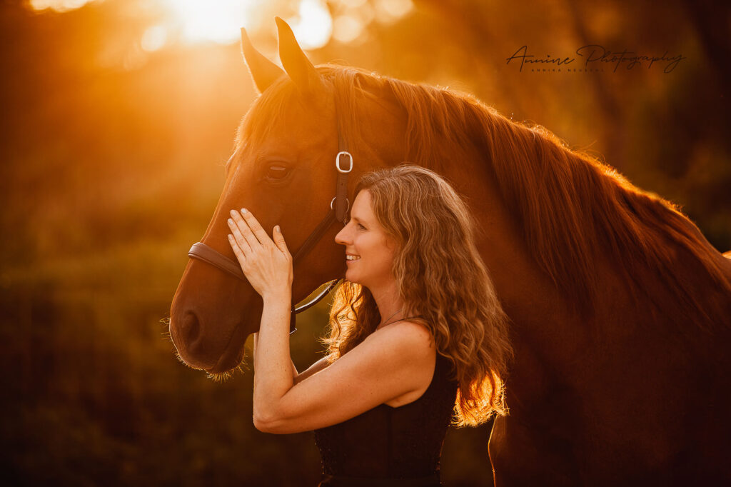 Daniela Landgraf steht neben einem schönen, braunen Pferd und streichelt dieses. Dabei lächelt Daniela Landgraf sympathisch und durch das Gegenlicht des Sonnenuntergangs entsteht ein insgesamt warmes, schönes Bild mit vielen Emotionen.