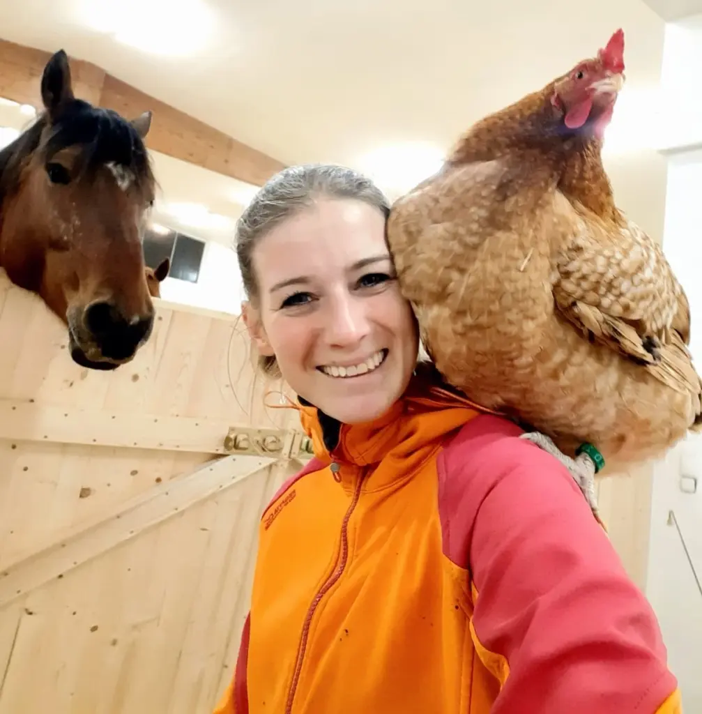 Ein Foto von Nina Schönrock, mit einem braunen Huhn auf ihrer Schulter, vor einem Pferdegehege posierend und in die Kamera lächelnd. Über das Gatter schaut ein braunes Pferd zu Nina.